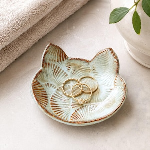 Puede incluir: Un plato de cerámica con forma de gato, esmalte verde claro y detalles texturizados en marrón. El plato presenta un patrón de abanico en toda su superficie y contiene tres anillos dorados finos, uno de ellos con una pequeña piedra.