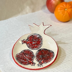 Peut inclure: Un plat en céramique en forme de grenade, intérieur blanc et bord rouge. Le plat présente trois sections de grenade, chacune avec un motif rouge et noir détaillé. Le haut du plat a une forme de couronne. Des pommes et une orange sont en arrière-plan.