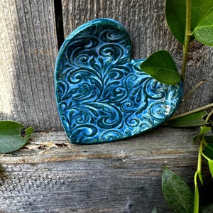 Puede incluir: Un plato de cerámica en forma de corazón azul verdoso con un patrón floral en relieve.
