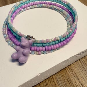 May include: A beaded wrap bracelet in shades of purple, blue, and white. The bracelet features a small, lavender gummy bear charm. The beads are small and round, creating a layered effect. The bracelet is displayed on a white surface.