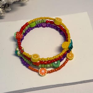 以下が含まれることがあります： レモン、オレンジ、キウイ、パイナップルなどのフルーツチャームが付いた、カラフルなビーズブレスレットのスタック。ブレスレットは赤、黄、紫、オレンジ色のビーズが特徴です。楽しくて夏らしいアクセサリー。