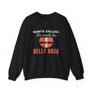 Pode incluir: Uma camisola preta de manga comprida com o texto SANTA CALLED. He wants his BELLY BACK em letras brancas, verdes e vermelhas. Um gráfico de um cinto de Pai Natal vermelho com uma fivela dourada está centrado entre as linhas.