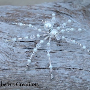 May include: A white beaded spider ornament with crystal accents. The spider has a silver wire frame and is decorated with white pearls and clear crystals.