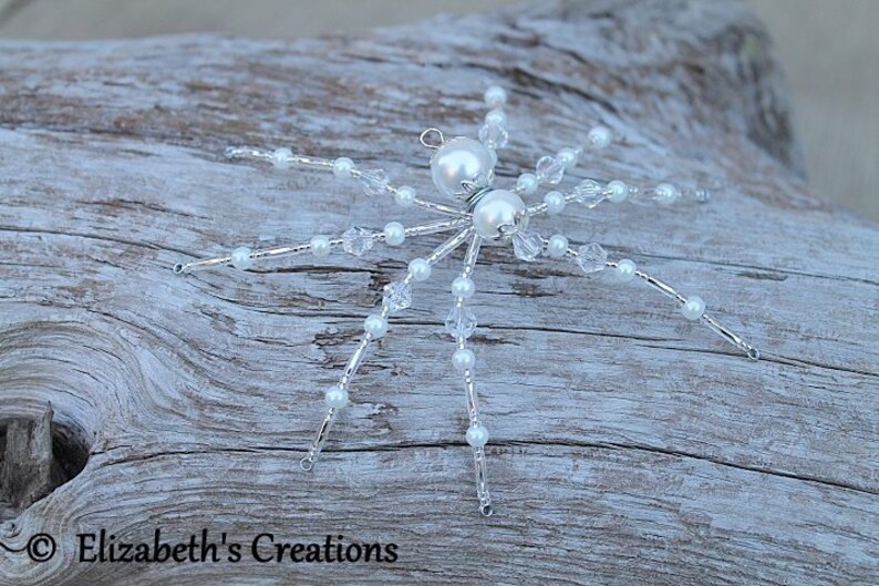 May include: A white beaded spider ornament with crystal accents. The spider has a silver wire frame and is decorated with white pearls and clear crystals.