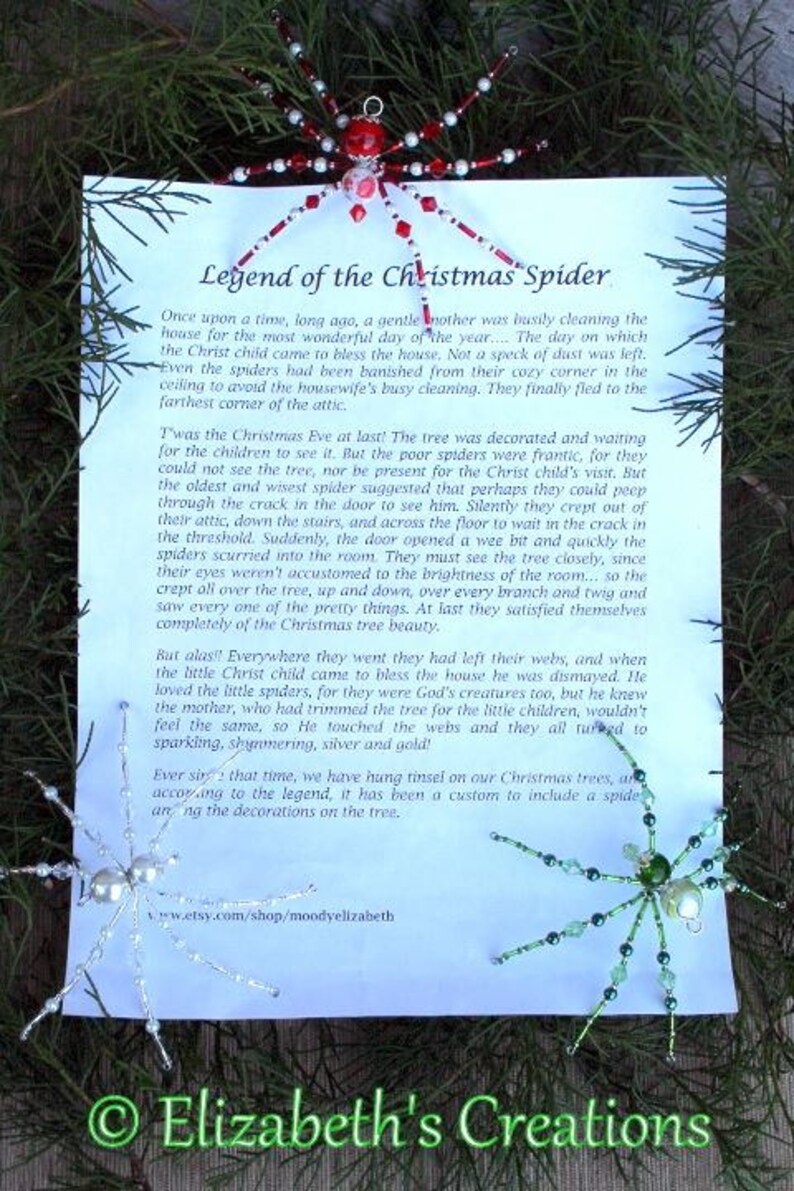 May include: A white card with blue text explaining the legend of the Christmas spider. The text describes how spiders were banished from a house being cleaned for Christmas Eve, but then crept back to see the decorated tree. The text says that the Christ child touched the spider webs, turning them to sparkling silver and gold. The card also shows a red and white beaded spider ornament and a green beaded spider ornament.