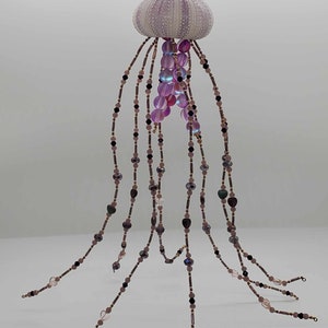 May include: A handcrafted jellyfish ornament featuring a lavender sea urchin shell body and beaded tentacles. The tentacles are strung with various shades of purple, pink, and black beads, creating a unique and decorative piece.