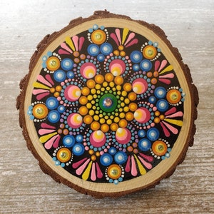 May include: A colorful mandala painted on a wooden slice. The mandala features a variety of colors, including pink, blue, yellow, and green. The mandala is surrounded by a black background.