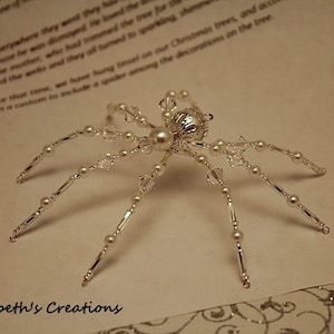 May include: A silver and white beaded spider ornament with a pearl in the center of the body. The spider has eight legs and is made of small beads and crystals.