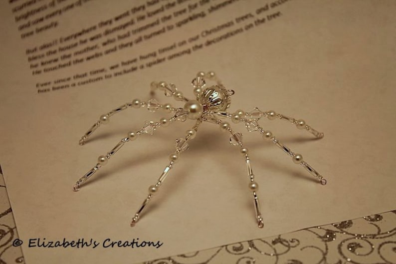 May include: A silver and white beaded spider ornament with a pearl in the center of the body. The spider has eight legs and is made of small beads and crystals.