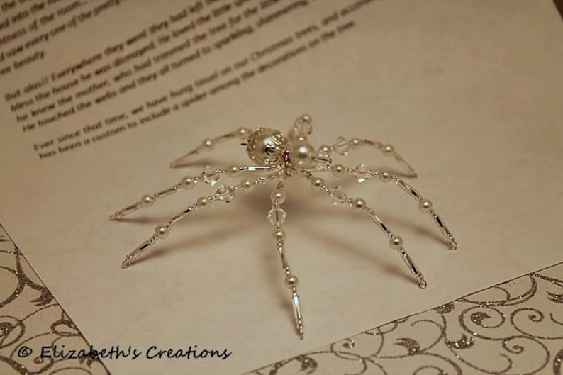 May include: A silver spider ornament with white pearl beads and crystal accents. The spider is on a white background with a decorative pattern.
