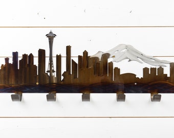 May include: Metal wall-mounted key holder shaped like the Seattle skyline, featuring the Space Needle, Mount Rainier, and several buildings. The holder has six hooks for hanging keys, coats, or other items.