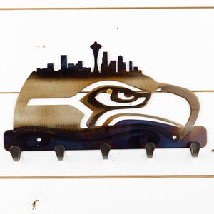 May include: Metal key holder featuring a silhouette of the Seattle skyline with the Space Needle and an eagle's head. The key holder has five hooks and a dark blue, purple, and gold finish.