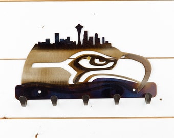 Könnte beinhalten: Metallischer Schlüsselhalter mit einer Silhouette der Skyline von Seattle mit dem Space Needle und einem Adlerkopf. Der Schlüsselhalter hat fünf Haken und eine dunkelblaue, violette und goldene Oberfläche.