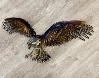May include: A metal eagle wall sculpture with iridescent blue, gold, and brown tones. The eagle is in flight with its wings spread wide and its talons extended.