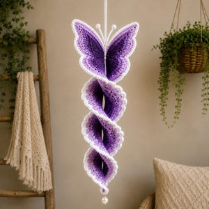 May include: A hanging crochet decoration featuring a purple and white butterfly at the top, transitioning into a long, spiraling, ombre-effect tail that fades from deep purple to light lavender, finished with a small white bead at the bottom.