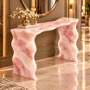 May include: A console table crafted from pink stone with a polished finish, featuring a smooth, wavy-edged top and two thick, undulating vertical supports. A white book titled The Kinfolk and a gold vase with white flowers sit on top.