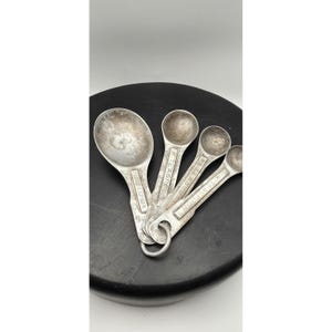 May include: A set of four silver-toned metal measuring spoons held together by a metal ring. The spoons are labeled 1 tablespoon, 1 teaspoon, 1/2 teaspoon, and 1/4 teaspoon. Each spoon has a worn, textured finish and a long handle.