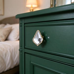 Pode incluir: Uma gaveta de cómoda em madeira verde-escura com um puxador de cristal facetado em forma de lágrima e rebordo em metal dourado. A frente da gaveta tem um acabamento mate suave com textura de grão de madeira visível e painel.