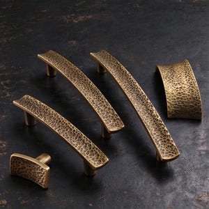 May include: A collection of five brass-toned cabinet hardware pieces, including four curved drawer pulls and one rectangular knob, all featuring a distinct hammered, textured surface with dark recessed detailing.