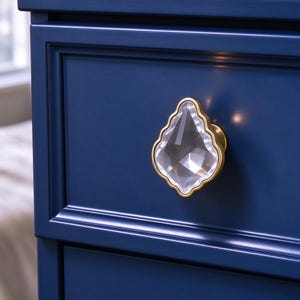 Könnte beinhalten: Ein Schubladenknopf mit einem klaren, facettierten, tropfenförmigen Kristall in einem goldfarbenen Metallrahmen. Der Knopf ist an einer tief dunkelblauen Schubladenfront mit einer rechteckigen Vertiefung montiert.