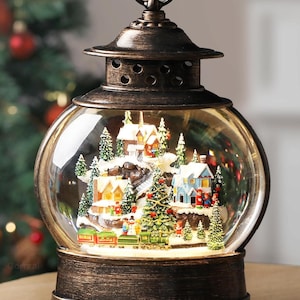 May include: A decorative lantern-shaped snow globe featuring a miniature winter village scene with houses, snow-covered pine trees, a train, and small figures. The base and top are finished in a dark, weathered bronze-toned metal.