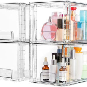 May include: A set of four stackable clear acrylic storage bins with grid-patterned sides. Two bins are empty, while the other two contain various skincare and cosmetic bottles, including dropper bottles, pump dispensers, and jars.