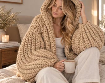 May include: A chunky knit beige hooded cardigan with long, voluminous sleeves and a thick, textured stitch pattern. The garment is worn over a simple white top, paired with loose-fitting white trousers, and held with a ceramic mug.