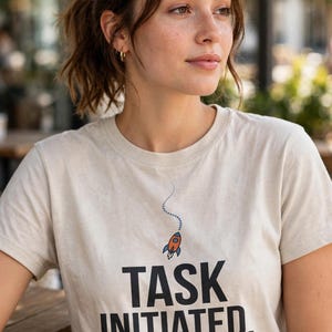 May include: A beige short-sleeved t-shirt featuring a small graphic of a rocket with a dotted trail above the bold black text that reads TASK INITIATED, INTEREST LOST. The shirt has a crew neckline and a standard fit.