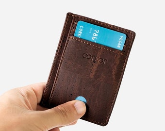 Puede incluir: Una cartera de corcho marrón con una tarjeta de crédito azul parcialmente insertada. La cartera tiene la palabra "corkor" grabada en relieve en la parte delantera. La tarjeta de crédito tiene el número "786" visible. La cartera se sostiene en una mano.