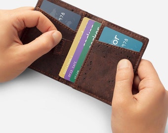 Puede incluir: Una cartera de corcho marrón con una banda elástica negra. La cartera está abierta y tiene varias tarjetas dentro, incluida una tarjeta azul con el texto "728" y una tarjeta amarilla con el texto "Sostenible, RFID Safe, Vegan".
