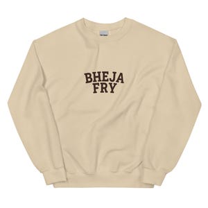 Op de afbeelding: Een zandkleurige sweater met ronde hals en de tekst BHEJA FRY in donkerbruine blokletters in het midden van de borst. Het kledingstuk heeft lange mouwen, een geribbelde kraag en geribbelde boorden, met een zichtbaar Gildan-label.