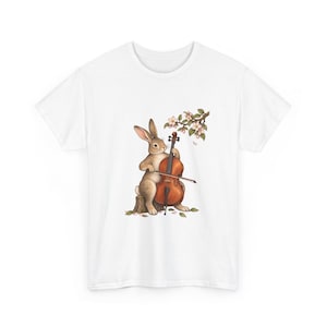 May include: A white t-shirt featuring a central graphic of a brown rabbit sitting on a tree stump, playing a wooden cello with a bow. Above the rabbit, a branch with light pink blossoms and green leaves hangs down, with scattered petals on the ground.