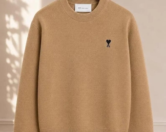 May include: A tan long-sleeve crewneck sweater featuring a ribbed collar, cuffs, and hem. The chest displays a small black embroidered logo consisting of a heart shape positioned above the capital letter A. A white brand label is visible at the inner neckline.