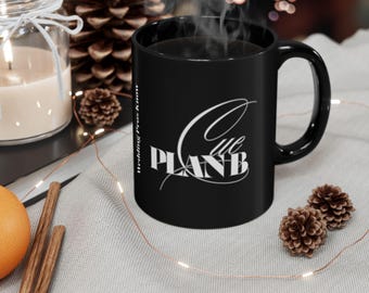 May include: A black ceramic mug featuring white text that reads "PLAN B" in a bold, blocky font, with the word "Cue" written in a cursive script above it. The vertical text "Wedding Pros Know" is printed along the side of the mug.