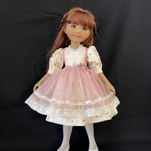 Könnte beinhalten: Eine Puppe mit langem rotbraunem Haar trägt ein cremefarbenes Kleid mit rosa Blumenmuster, einen rosa Tüllüberwurf mit Spitzenbesatz, weiße Strumpfhosen und weiße Schuhe. Das Kleid hat Puffärmel und eine kleine rosa Rose an der Taille.
