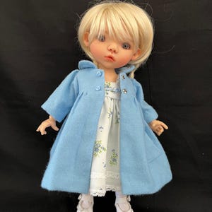 Puede incluir: Muñeca con cabello rubio y ojos azules que lleva un abrigo azul claro sobre un vestido blanco con estampado floral. La muñeca también lleva zapatos blancos. El abrigo tiene tres botones y un cuello. La muñeca mide unos 30 cm de altura.