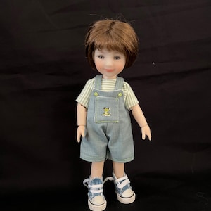 May include: A doll with short brown hair wearing a light green and white striped short-sleeve shirt under light blue denim overalls featuring a small frog graphic on the chest pocket. The doll wears blue and white canvas sneakers with white laces.