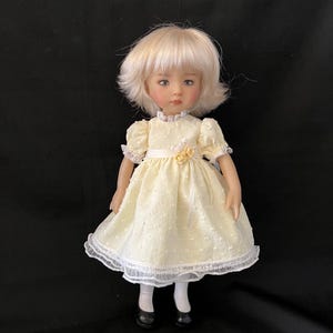 Puede incluir: Muñeca de porcelana con cabello rubio y ojos azules, vestida con un vestido amarillo pálido con ribete de encaje blanco. El vestido tiene una cinta en la cintura y un pequeño adorno floral. La muñeca lleva calcetines blancos y zapatos negros.