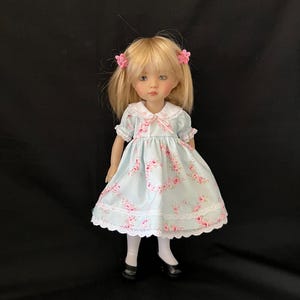 May include: A doll with blonde hair styled in pigtails, adorned with pink flower hair clips. The doll wears a light blue dress with a pink rose pattern, white lace trim, and black shoes. The doll's dress has a white collar and a pink bow.