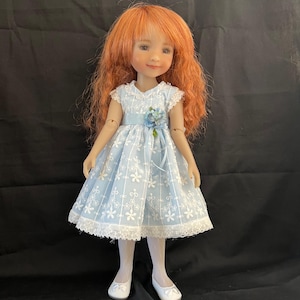 May include: A doll with red hair wearing a light blue dress with white floral patterns and lace trim. The dress has a blue ribbon and a blue flower accent. The doll is wearing white tights and white shoes. The doll is standing in front of a black background.
