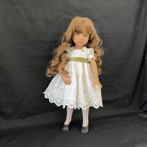 Puede incluir: Una muñeca con cabello largo y ondulado castaño y ojos azules, vestida con un vestido blanco con bordado floral y una cinta verde. La muñeca también lleva medias blancas y zapatos negros. La muñeca está de pie frente a un fondo negro.