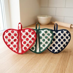 May include: Three heart-shaped quilted pot holders featuring a white background with polka dots in red, green, and navy blue. Each pot holder has a matching fabric loop at the top for hanging and is finished with a coordinating fabric trim.