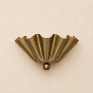 May include: A wall-mounted light fixture featuring a wide, semi-circular brass shade with deep, vertical fluted pleats. The base of the shade is accented with a single, centered brass sphere, creating a structured and metallic appearance.