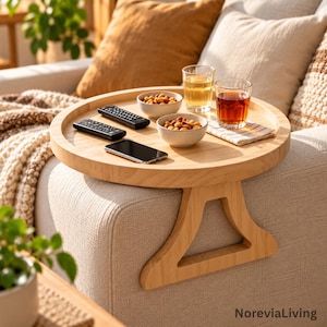 Könnte beinhalten: Ein rundes, helles Holztablett für Sofalehnen mit einer flachen Oberfläche und einem seitlichen Stützelement. Auf dem Tablett liegen zwei schwarze Fernbedienungen, ein schwarzes Smartphone, zwei kleine Schalen mit Nüssen und zwei Gläser.