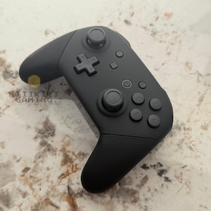 May include: A matte black wireless video game controller featuring two analogue sticks, a directional pad, and four action buttons labelled A, B, X, and Y. The controller includes a central home button and a small logo reading KIIKINI GAMING.