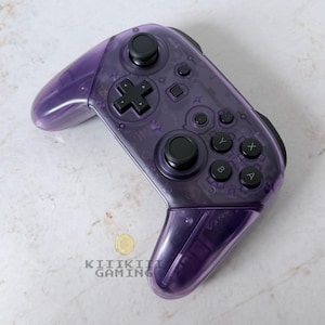 May include: A wireless video game controller with a translucent purple plastic shell. The device features black analog sticks, a directional pad, and action buttons labeled A, B, X, and Y. The text KIIIKIII GAMING is printed in black near the bottom.