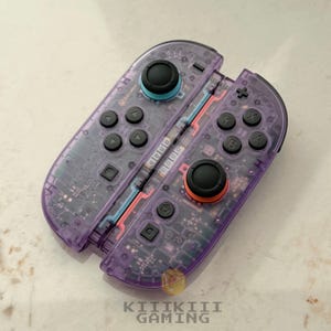 May include: A pair of translucent purple gaming controllers featuring black buttons, a light blue joystick cap, and an orange joystick cap. The internal circuitry is visible through the clear casing. The text KIIKIII GAMING is printed at the bottom.