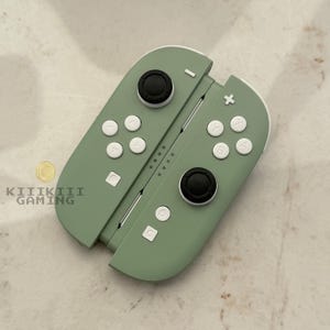 May include: A pair of sage green gaming controllers featuring black analog sticks and white buttons labeled with symbols including a plus sign, a minus sign, and letters A, B, X, and Y. The text KIIIKIII GAMING is printed in black on the left side.