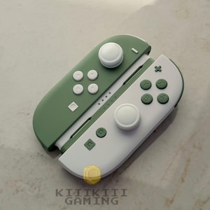 May include: A pair of gaming controllers featuring a two-tone design. The left controller is sage green with white buttons and a white joystick. The right controller is white with sage green buttons and a white joystick. The text KIIIKIII GAMING is visible.