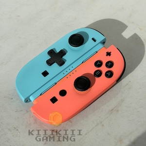 May include: A light blue and coral video game controller set featuring black analog sticks, a black directional pad, and black action buttons labeled A, B, X, and Y. The text KIIKIII GAMING is printed in a pixelated font at the bottom.